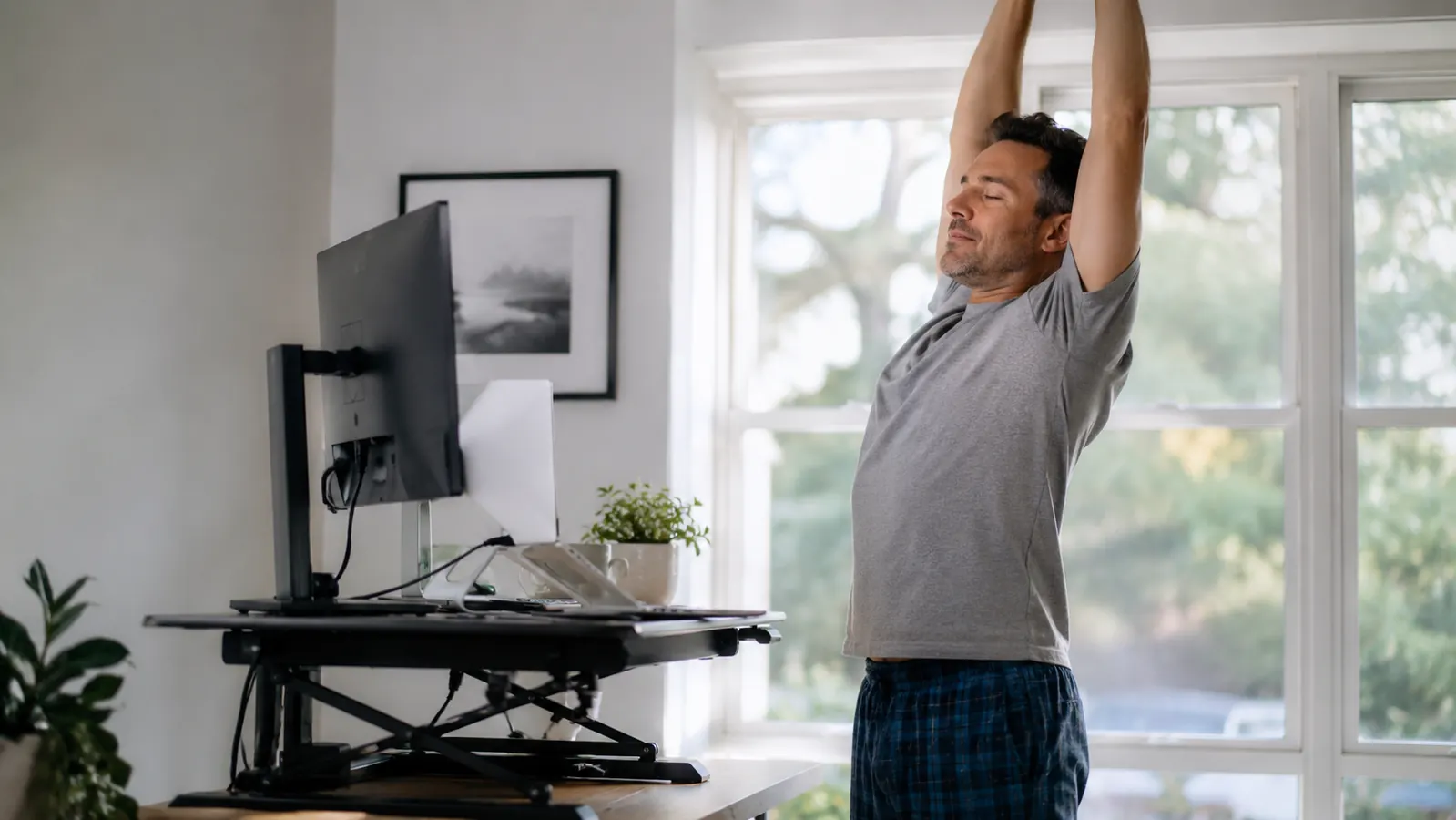Remote worker standing at raised desk converter, stretching arms overhead during a break, bright natural light